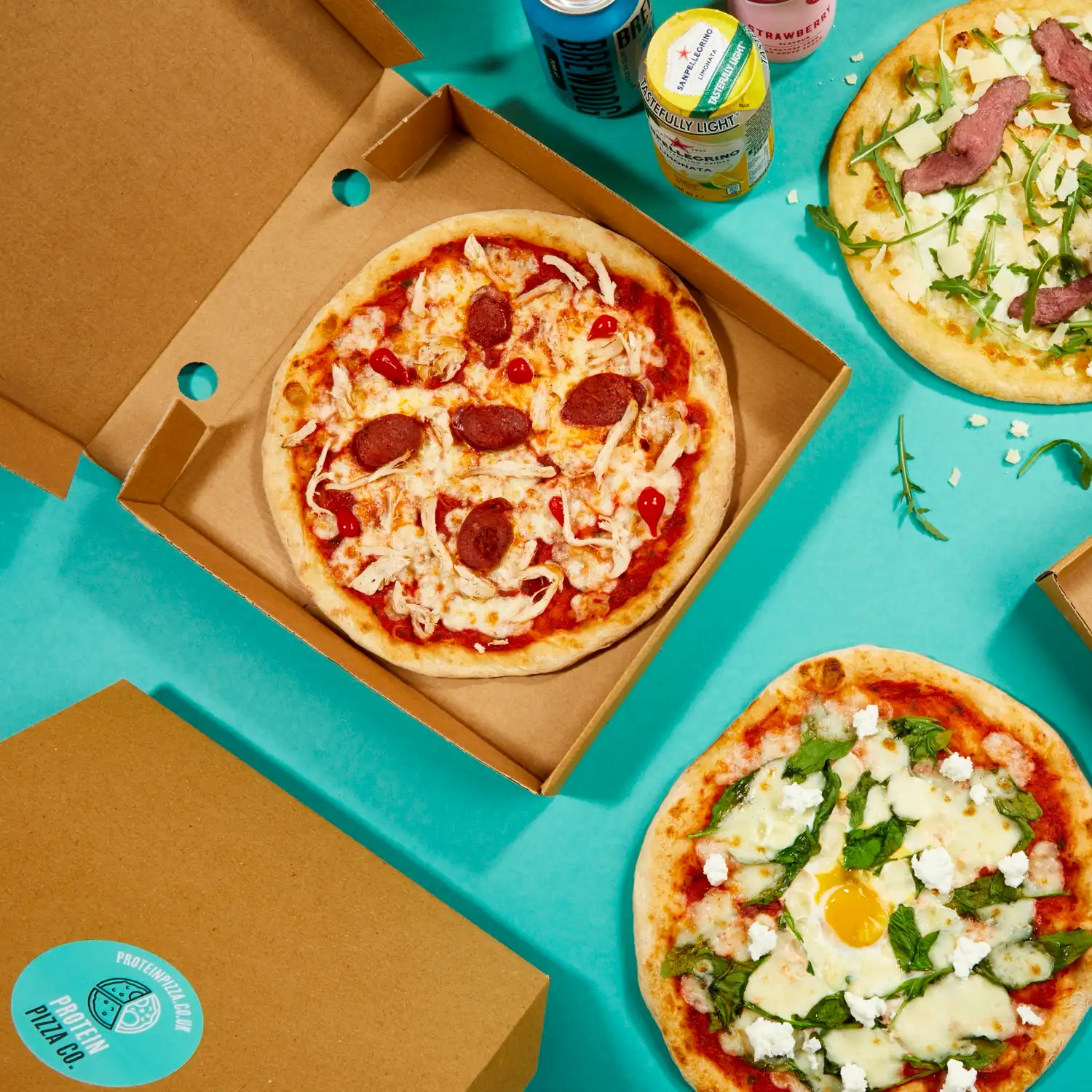 Protein Pizza Co. boxes and pizzas arranged on a teal background with canned drinks