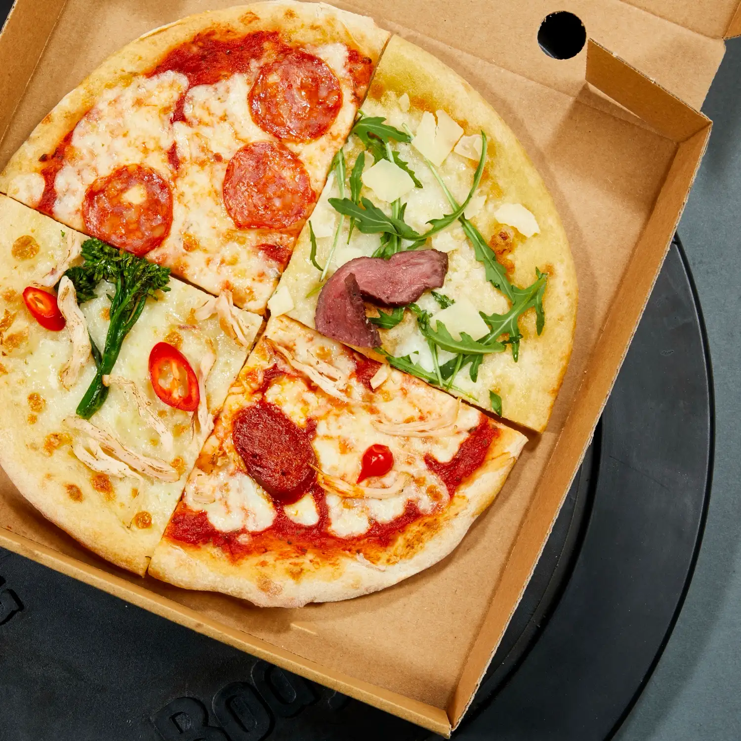 A quartered Protein Pizza Co. pizza in its box, each quarter a different topping, resting on a weight plate