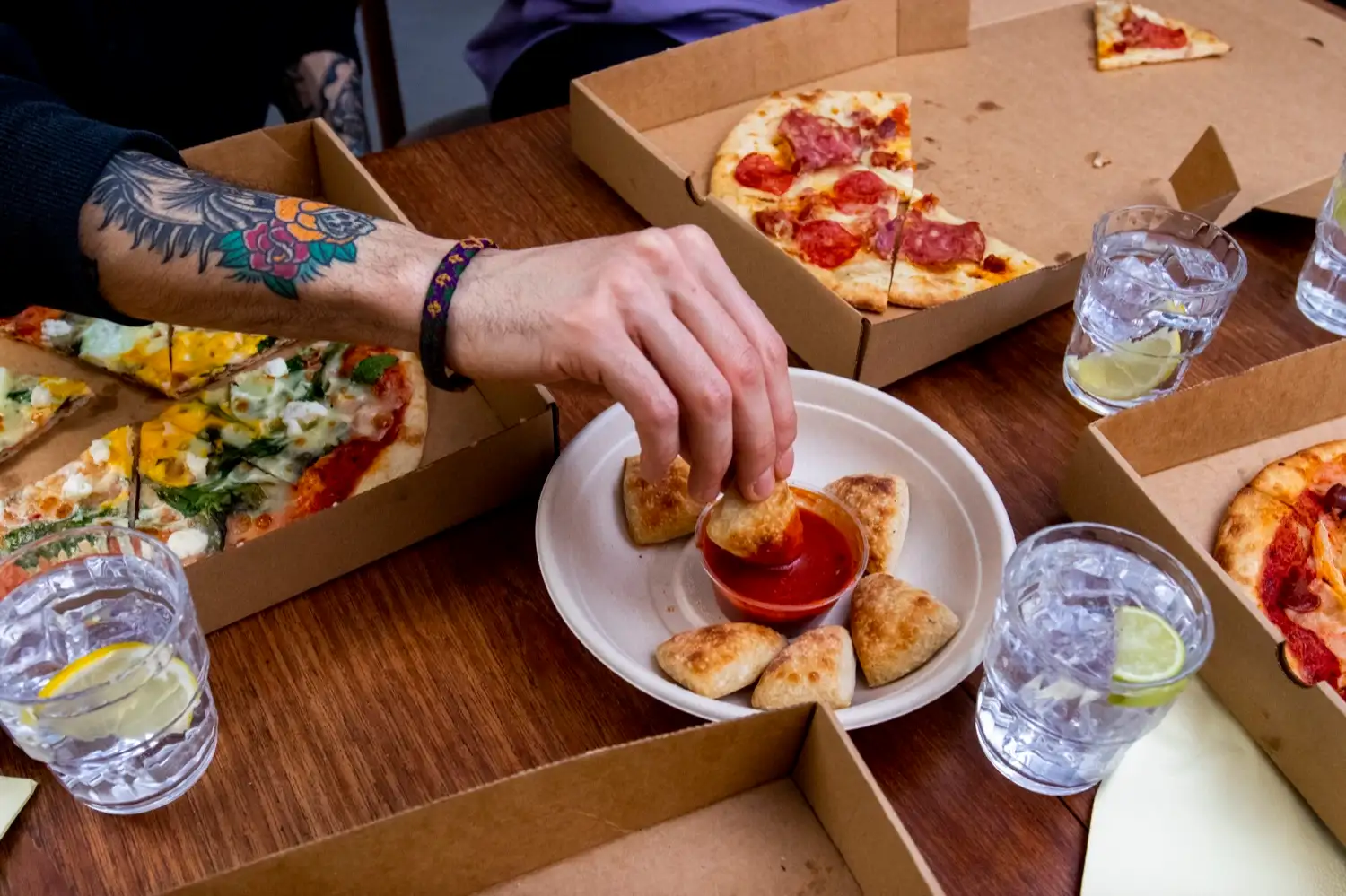 Hands reaching for protein dough balls and dip across a table of pizzas